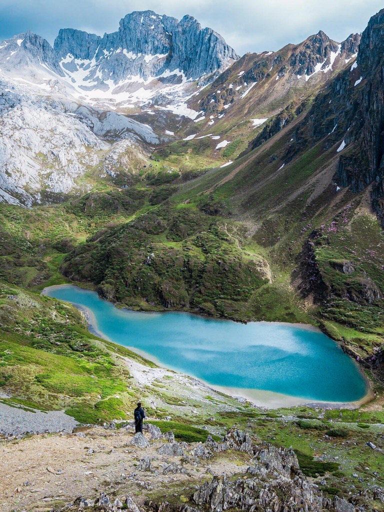 In Abujicuo Shangri-La a hiker stands on rocky terrain overlooking a turquoise lake surrounded by green hills and snowy mountains under a cloudy sky. Serene scene.