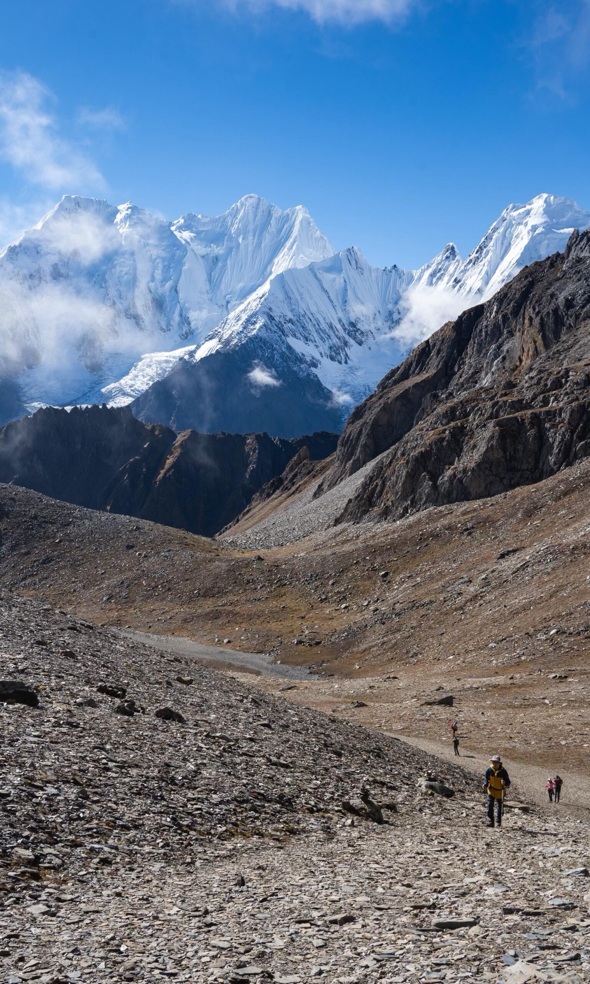 In Meili North Slope Shangri-La, hikers trek a rocky mountain trail with snowy peaks in the background under a clear blue sky. Distant figures show exploration mood.