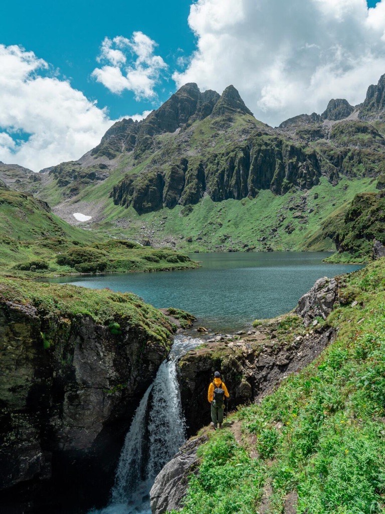 In Nanjiluo, Shangri-La, a hiker in a yellow jacket stands by a waterfall and lake, set against green mountains under a partly cloudy blue sky. Mood is serene and adventurous.