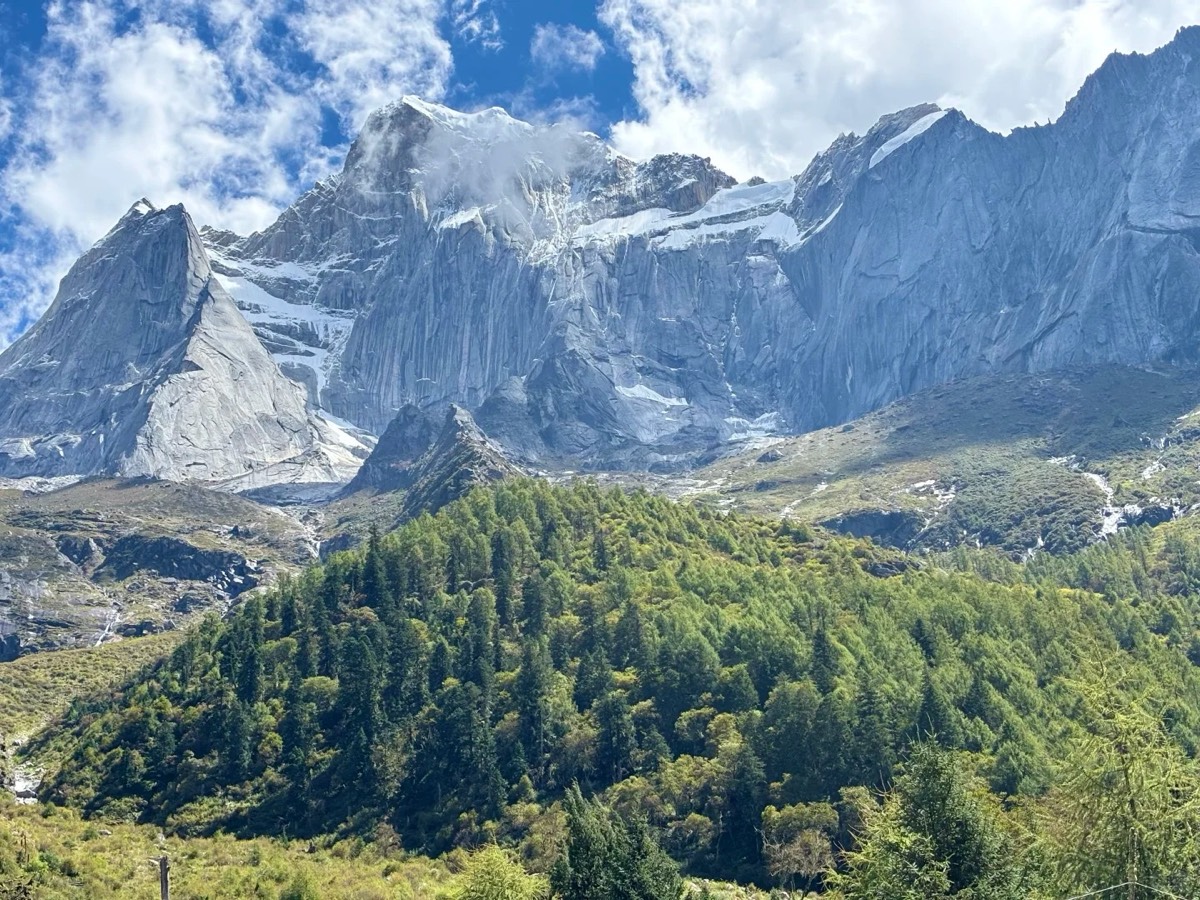 A close-up of a massive, rugged gray rock face with patches of snow and ice, rising sharply above a 