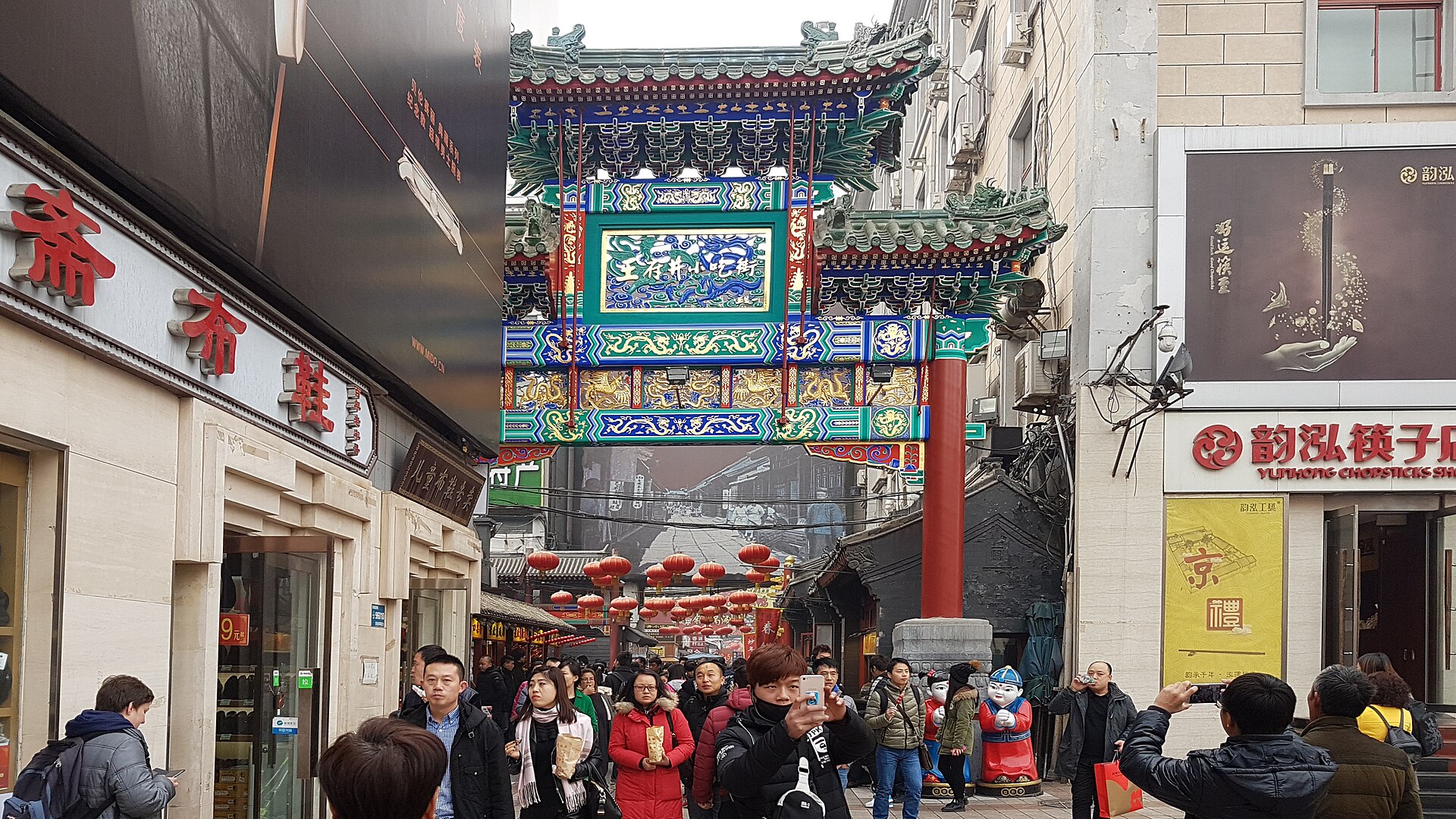 The brightly painted paifang archway marking the entrance to Wangfujing Snack Street (王府井小吃街), red lanterns over a lively crowd, Beijing