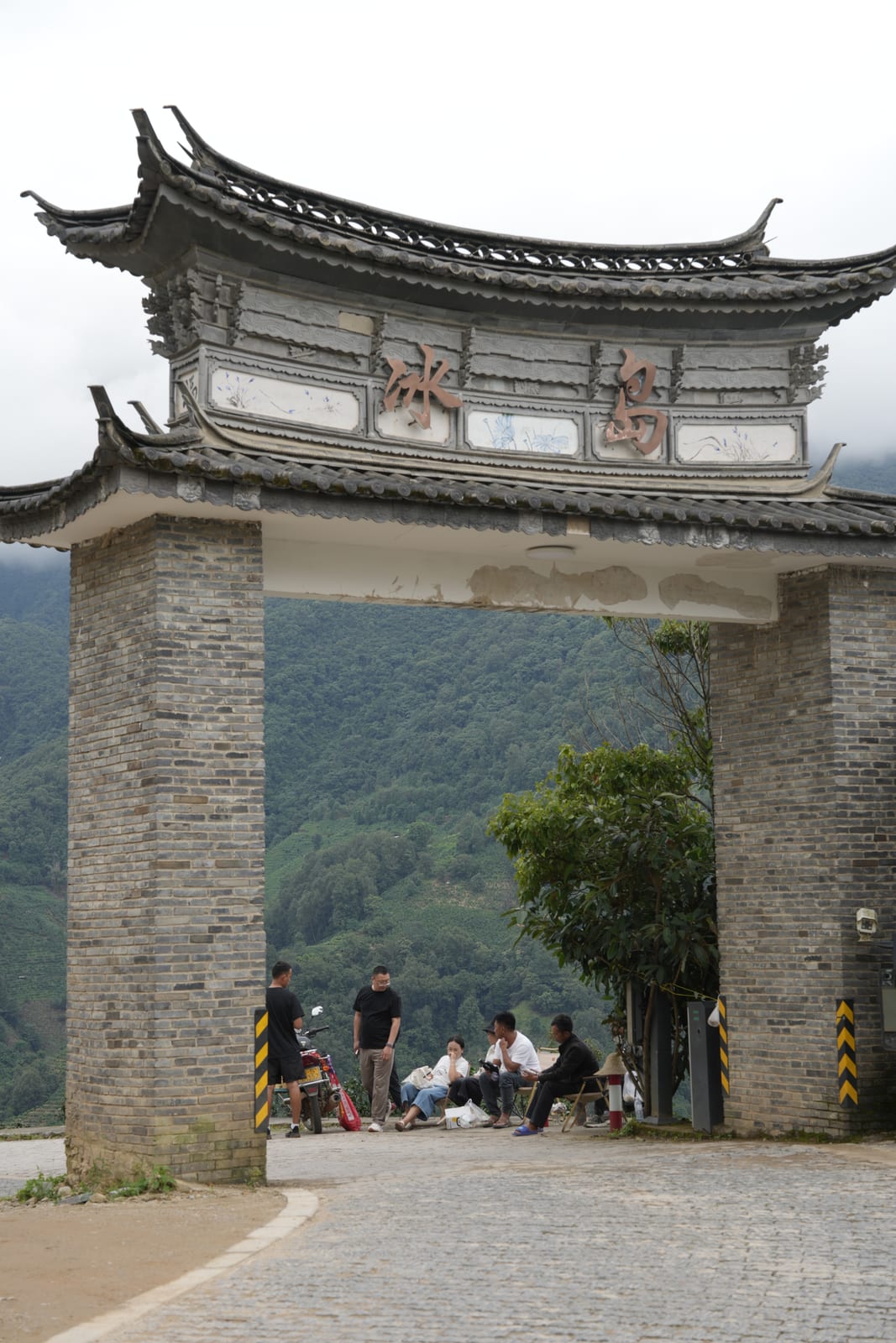 Travellers at the carved stone-and-tile village gateway of Bingdao Laozhai, tea mountains beyond