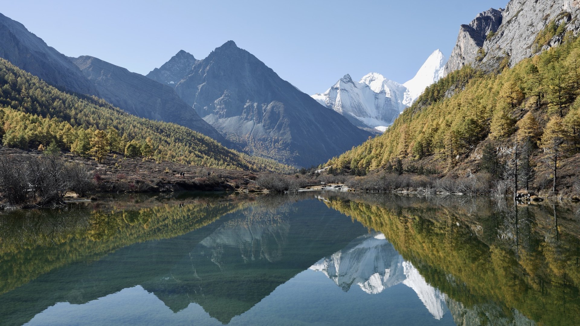 A still lake bend in Bipeng Valley mirrors jagged grey peaks, a snow dome and autumn larch
