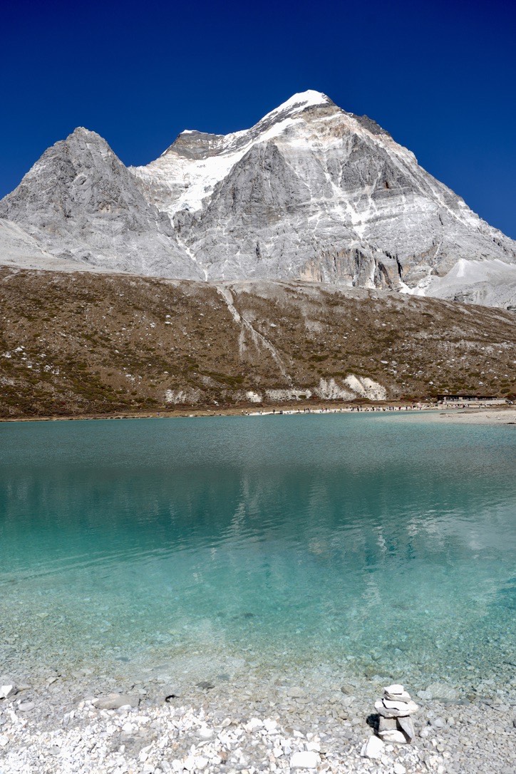 Turquoise glacial waters of Milk Lake beneath a snow-dusted grey peak under deep blue sky on the Yading long kora.
