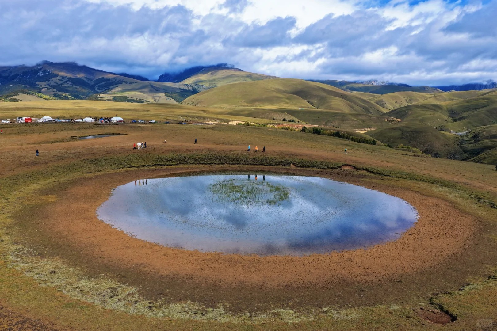 Aerial over the Eye of Genie — the oval pool holds cloud and sky, trekker camp in the meadow beyond