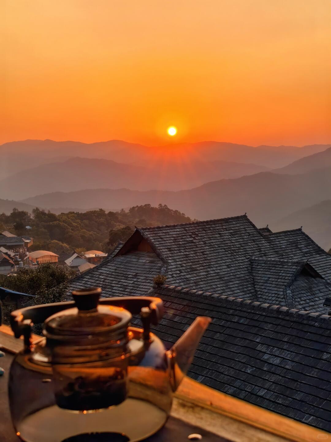 Glass teapot brewing tea on a wooden tray with the sun rising over Mangjing village rooftops and layered Jingmai ridges — last tea before the morning departure