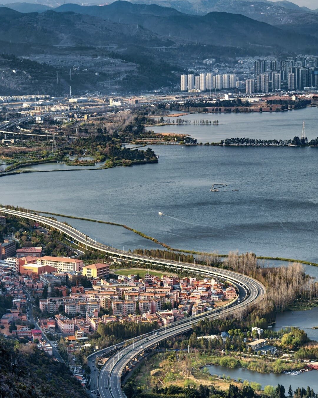 Aerial view over Kunming's Dianchi lake with a sweeping highway, lakeside town and high-rises backed by mountains