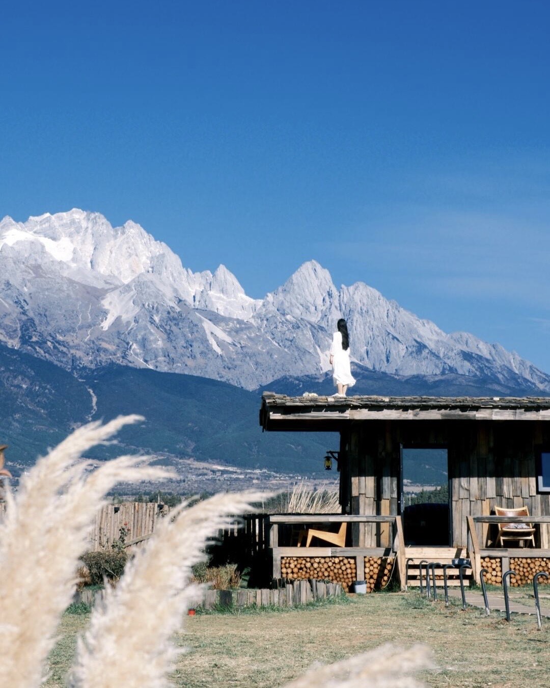 Woman in white on a wooden cabin roof facing the snow-capped ridges of Jade Dragon Snow Mountain near Baisha.