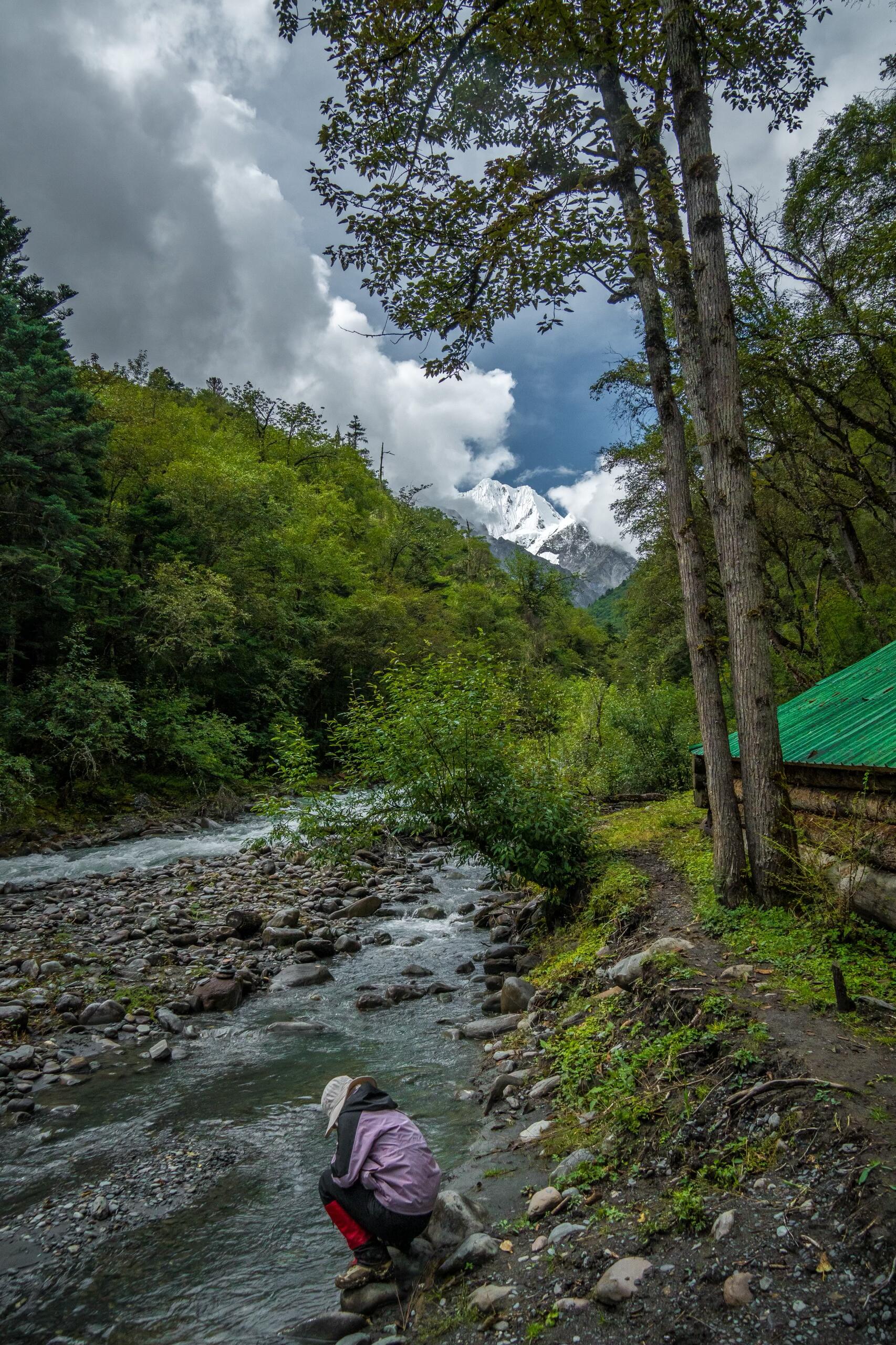 Riverside rest stop in lush forest with snow peaks through the clouds — Day 3 trek-in