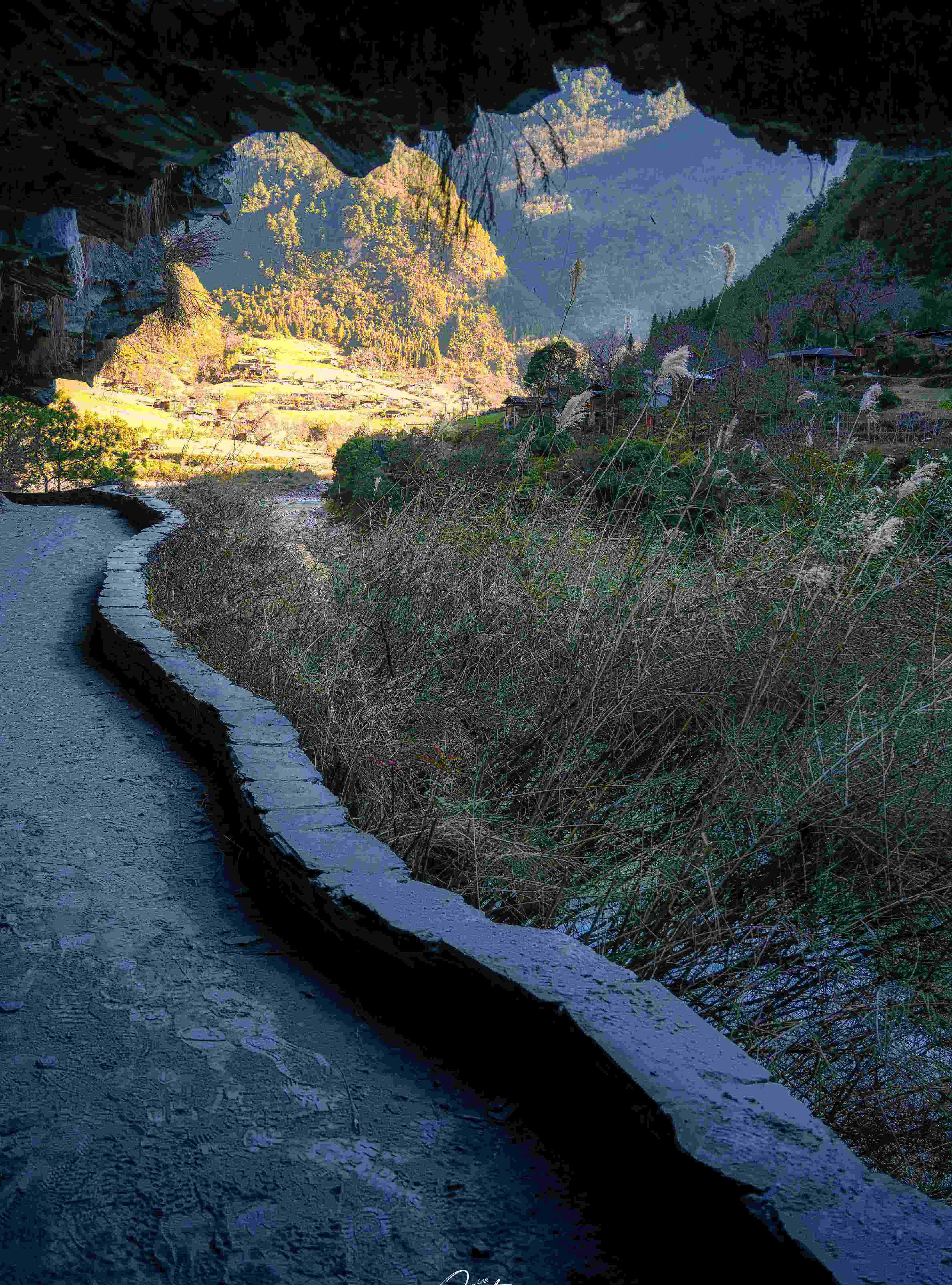 A stone trail emerges from a rock overhang onto sunlit Nujiang terraces beyond