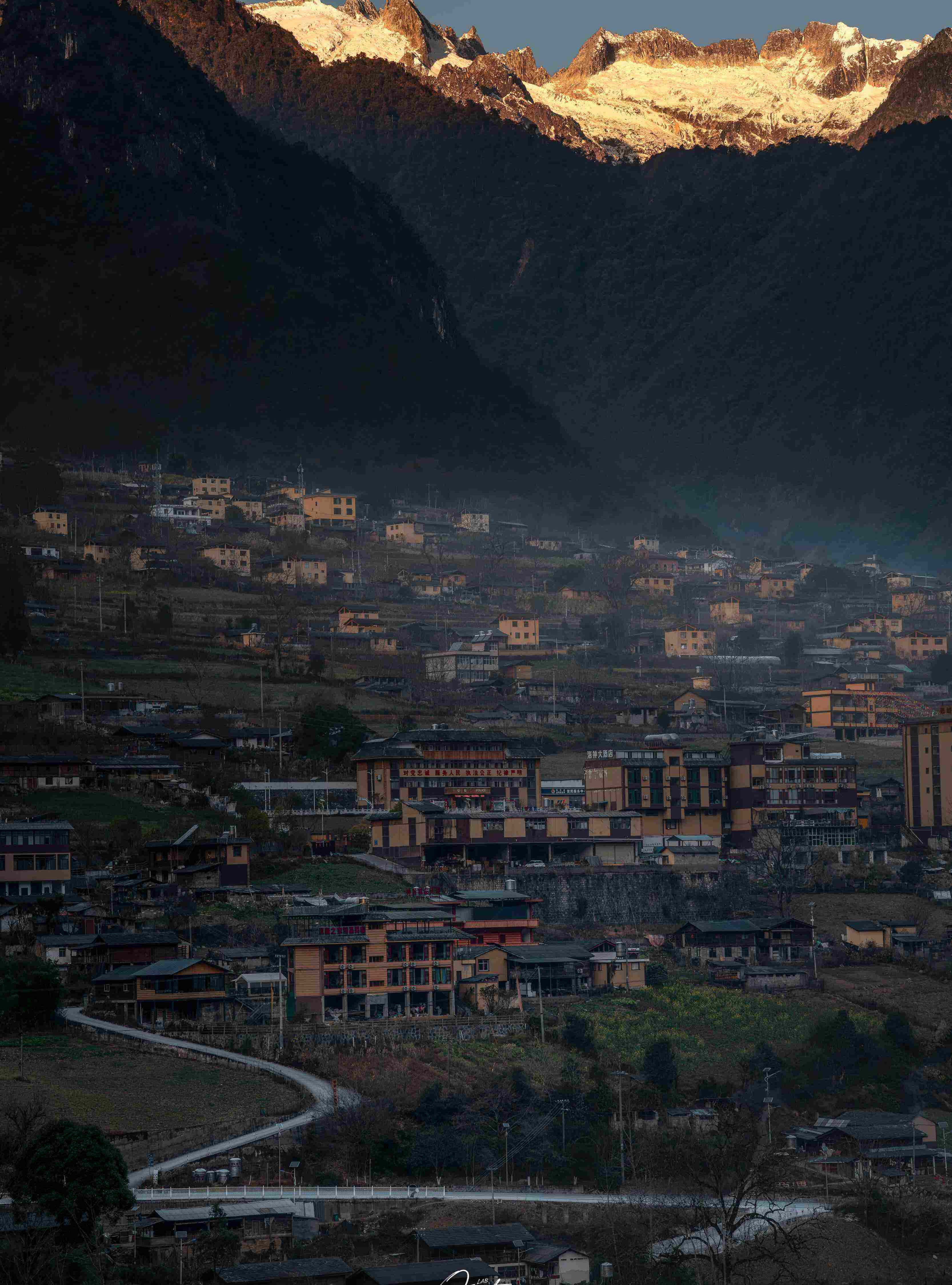 Nujiang valley village in alpenglow — snow peaks ignited gold above tile-roof homes in shadow