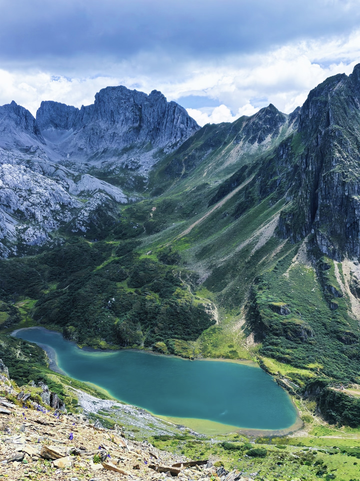 Hiker on rocky terrain overlooking the turquoise Abujicuo alpine lake ringed by snow-spotted ridges above Shangri-La — China's Last Frontier loop