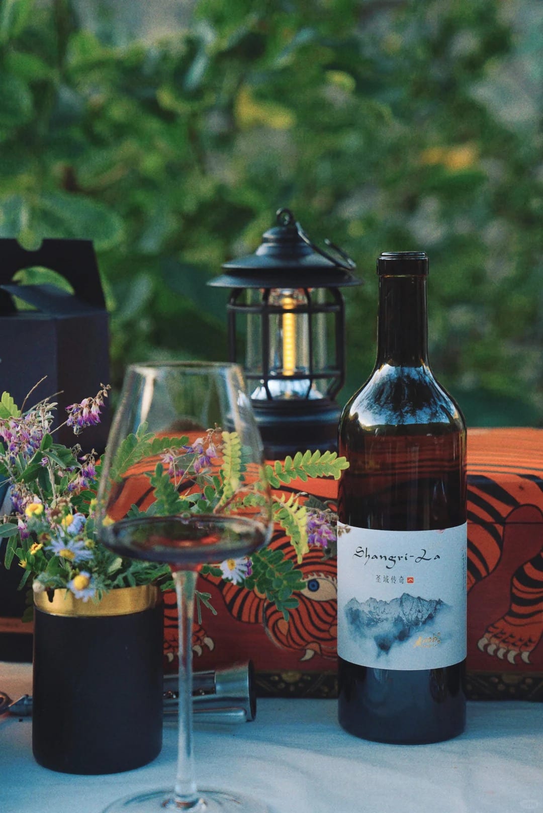 A glass of red wine, a lantern and a bottle on a table set among the vines in the Shangri-La wine region — Shangri-La Wine Journey