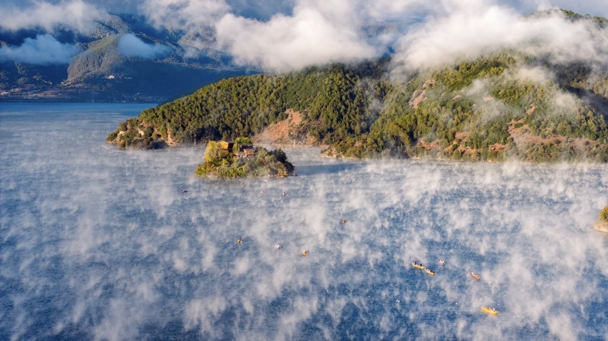 Morning mist drifts over Lugu Lake as canoes glide past its forested island