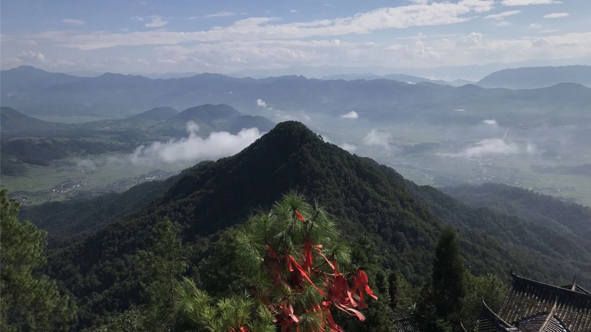 Forested volcanic cone rising above Tengchong from a ribbon-strung summit viewpoint — Yunnan slow culture tour