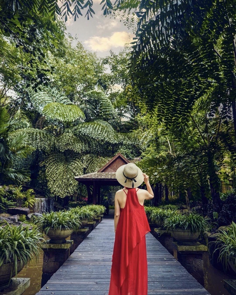 Woman in a red dress and straw hat walking a boardwalk through lush ferns and potted plants toward a pavilion.
