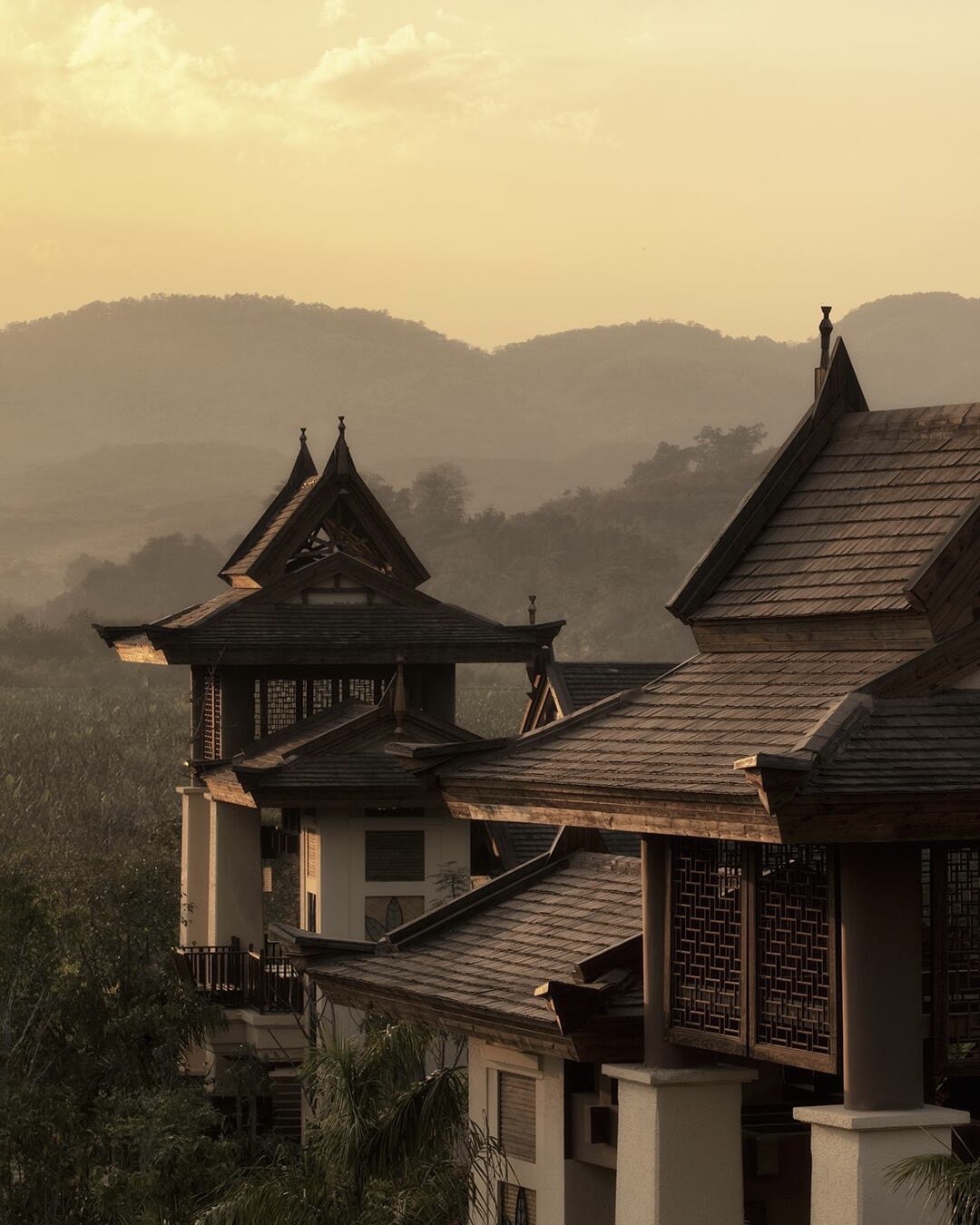Tiered timber pavilion roofs with carved lattice screens against misty forested hills at golden sunset, Anantara Xishuangbanna.