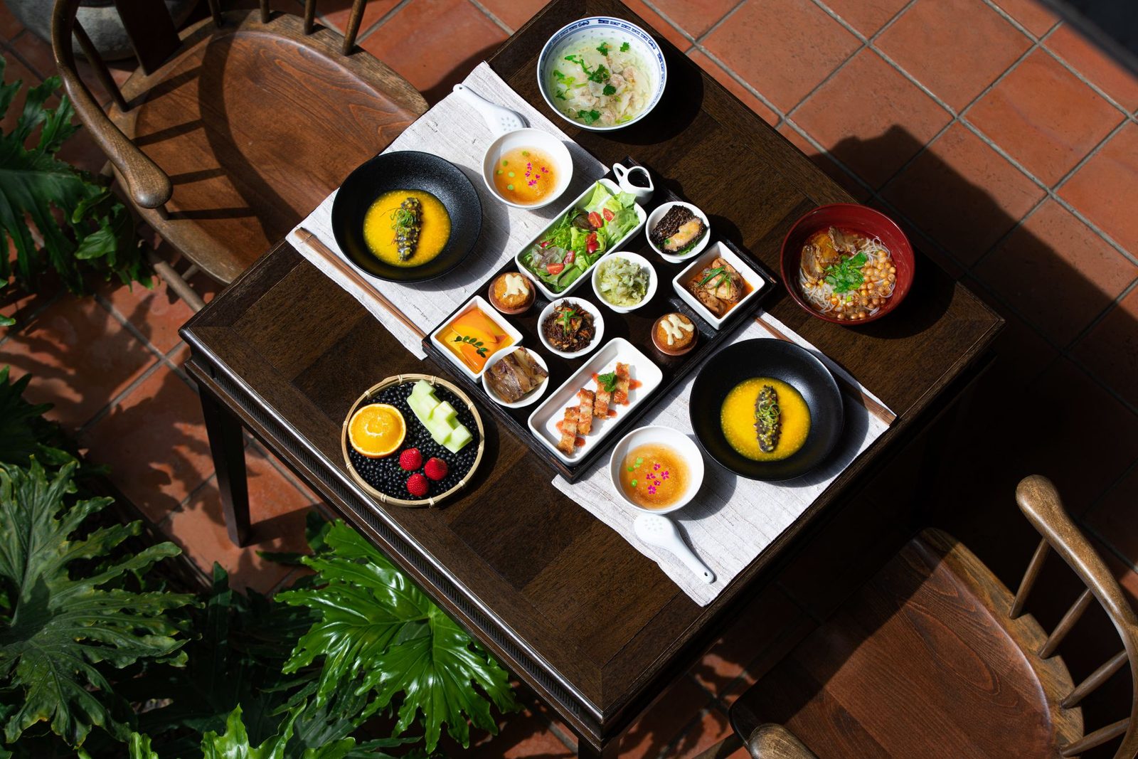 Breakfast of small Yunnan dishes, fruit and noodles laid out on a courtyard table among the plants.
