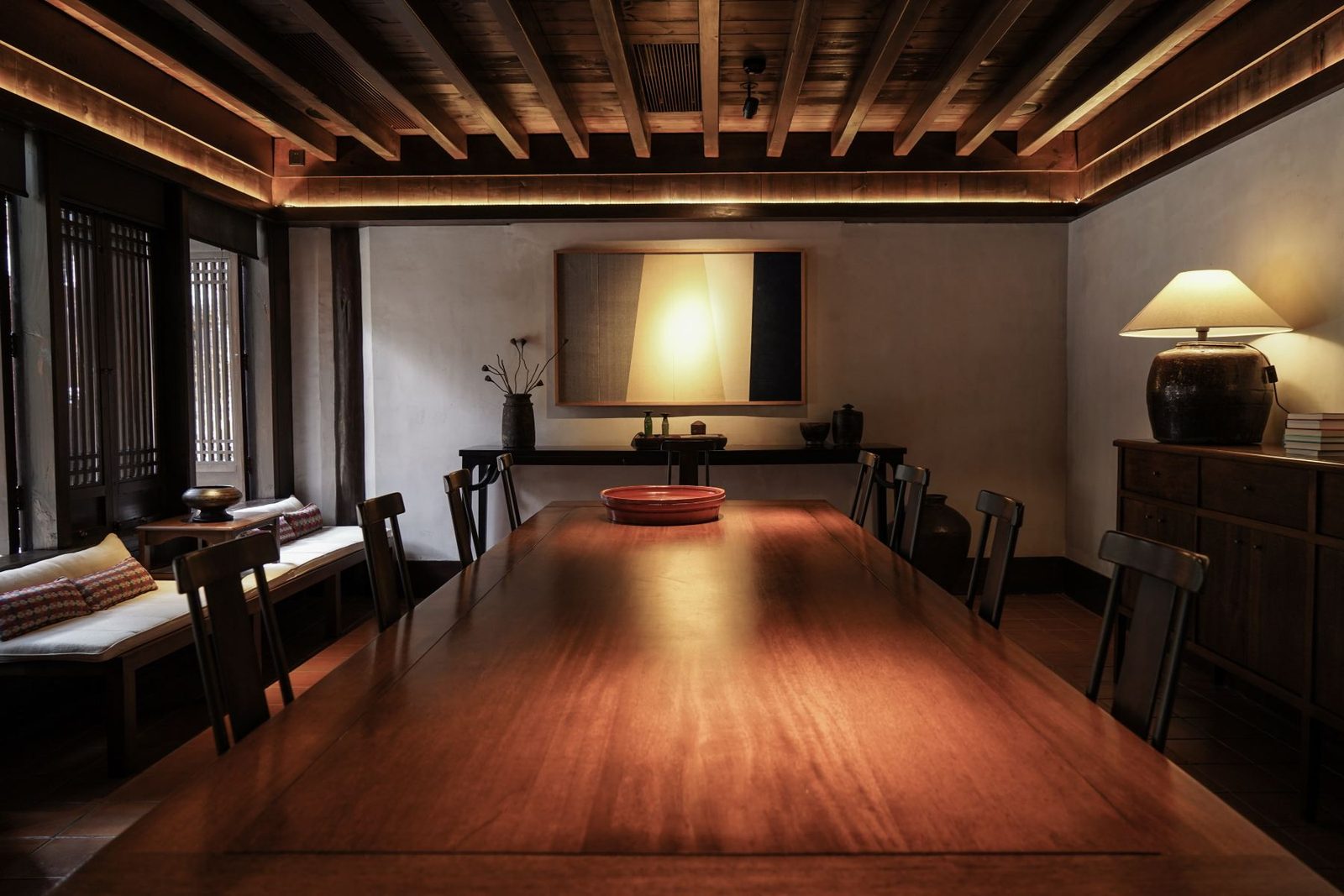 Long polished-wood table under a beamed ceiling, a lamp and daybed along the wall.