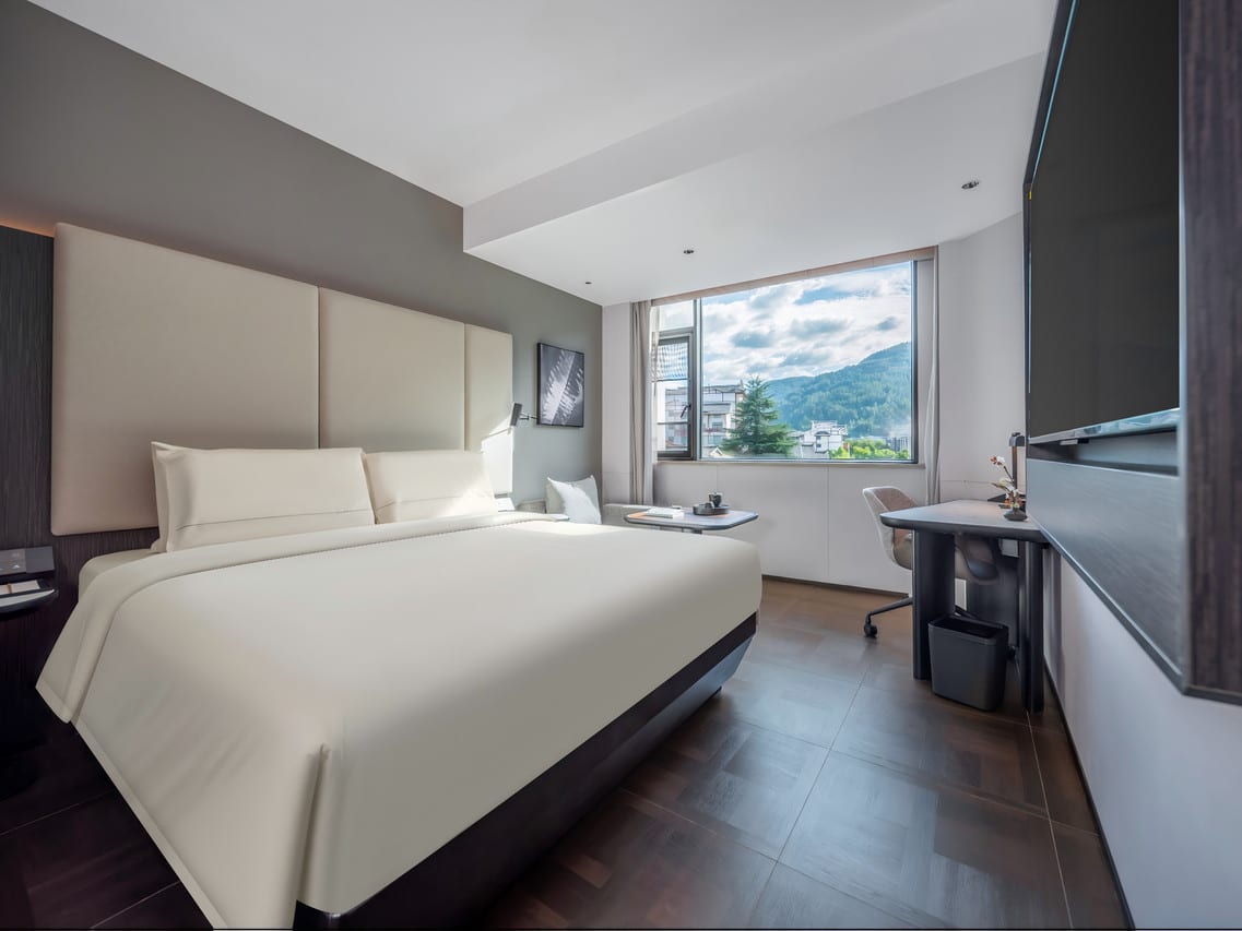 Atour Hotel, Zhangjiajie Forest Park Gate — a room with a large double bed against tall upholstered headboard panels, the window framing green hills above white-walled houses
