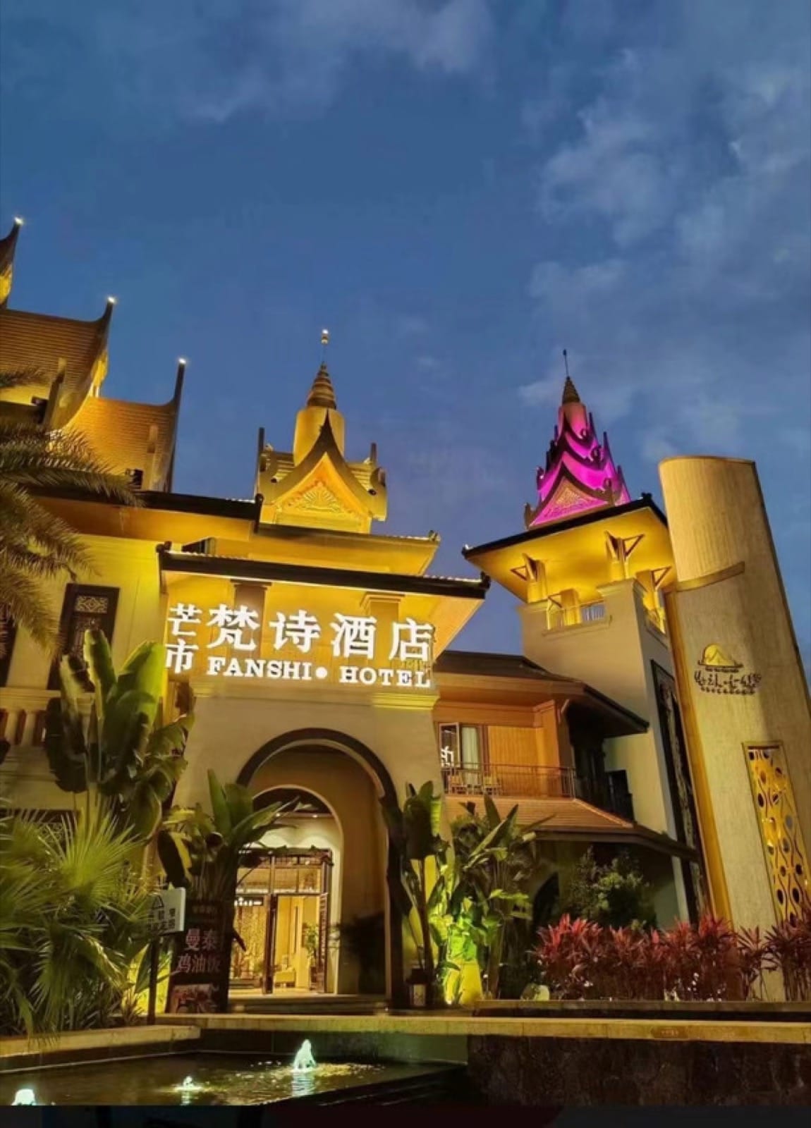 Fanshi Hotel — gold-spired Dai towers lit at dusk above an arched gateway and a courtyard fountain, banana palms in the foreground, Mangshi