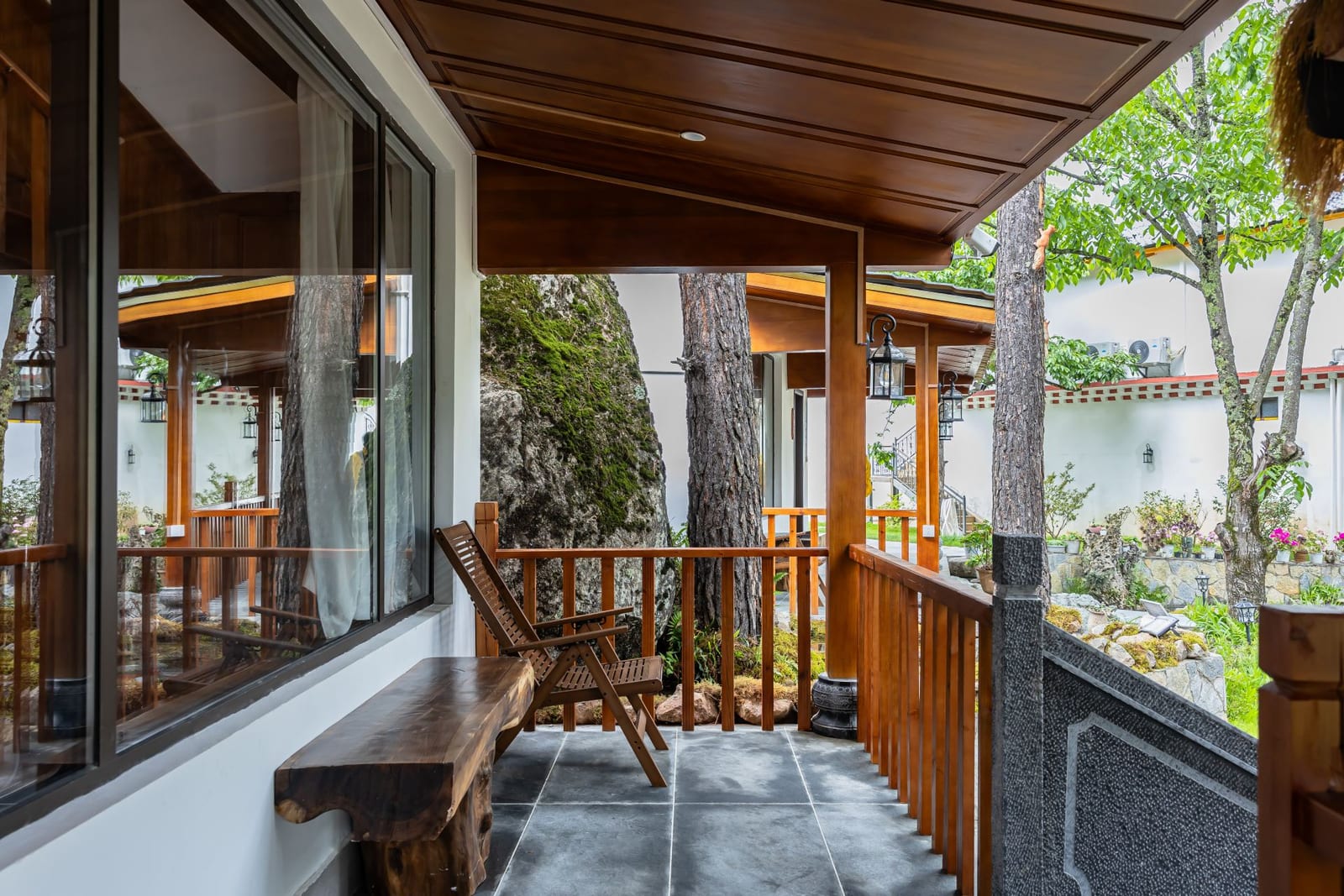 Jilin Mountain Lodge — a timber balcony with a bench and railing outside a room, pine trunks and the garden wall beyond