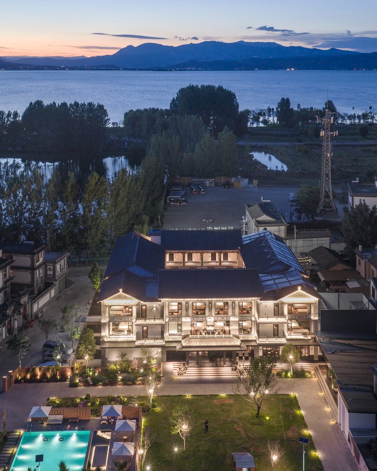 Kasion Erhai from above at dusk — the lit Bai courtyard wings and pool, with Erhai and the Cang Mountain ridge behind, Xizhou