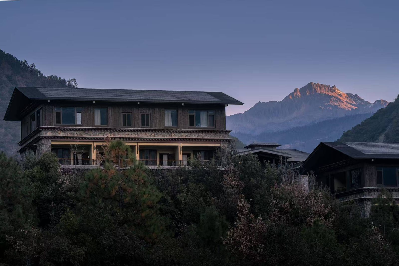 Songtsam Lodge Cizhong — the stone-and-timber lodge buildings in evening light, a snow peak catching the last of the sun behind them