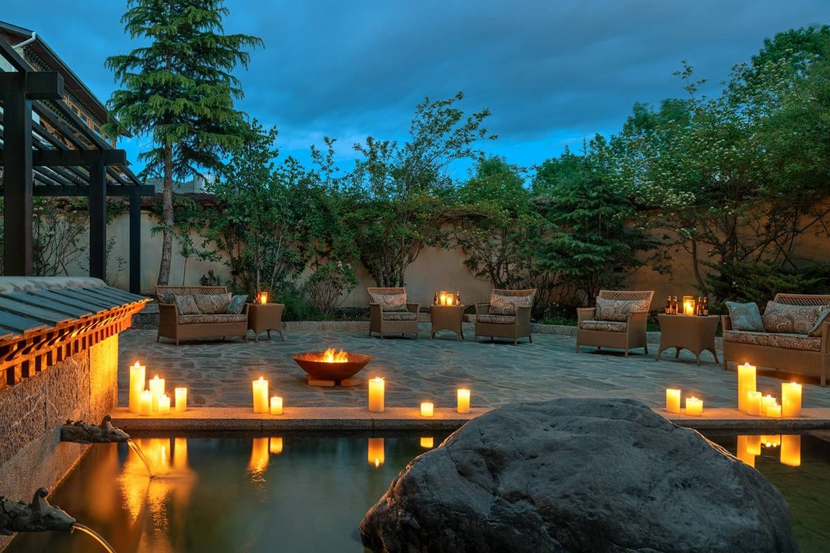 Songtsam Lodge Shangri-La — a walled stone terrace at dusk, wicker armchairs around a fire bowl and candles lining a pool fed by spouts in the wall