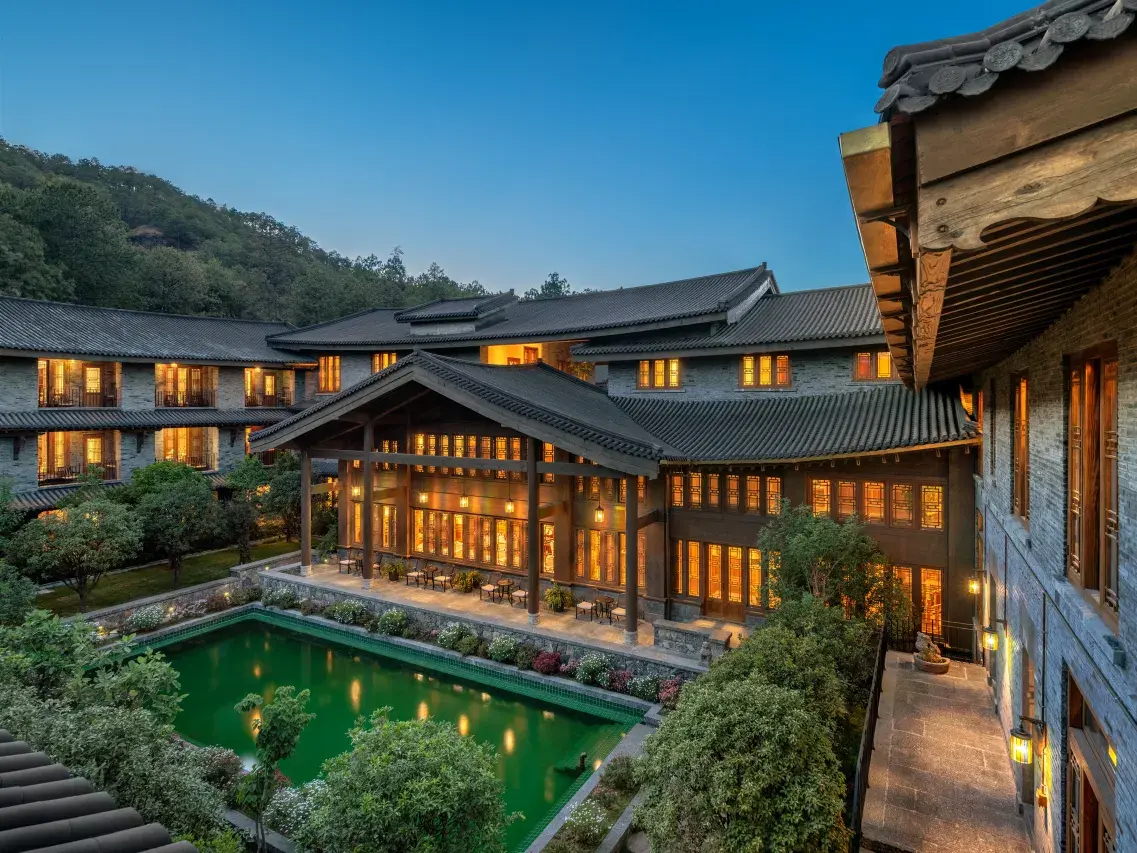 Illuminated grey-tiled pavilion and guest wings ringing a green courtyard pool at dusk.