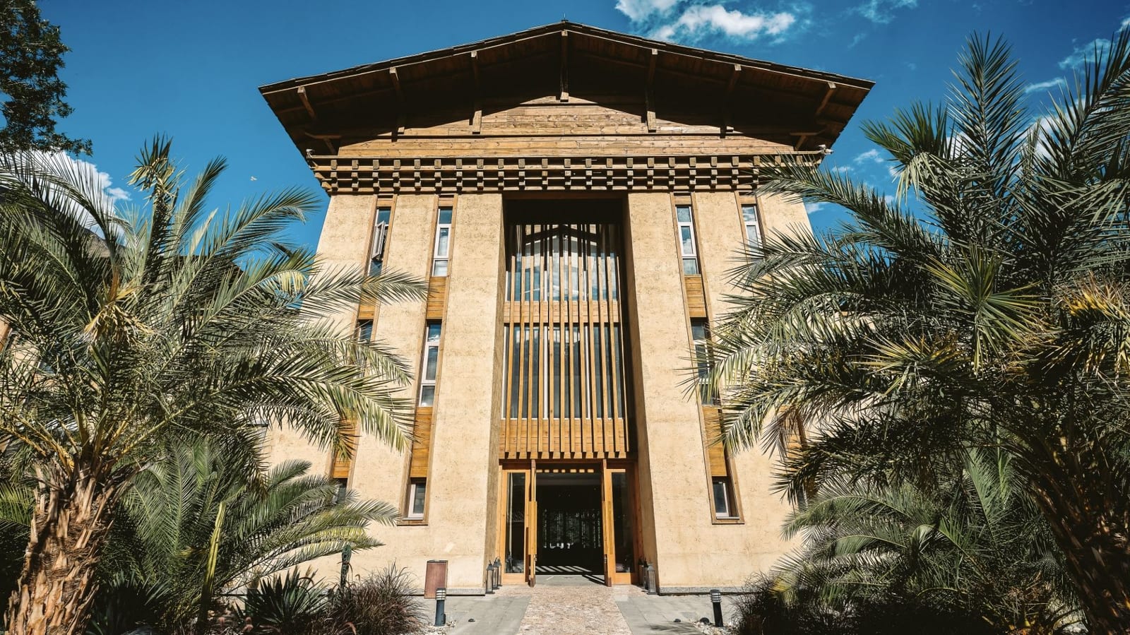 Tea Horse Road Benzilan — the rammed-earth tower and tiered timber eaves of the entrance façade between palm trees under a blue sky