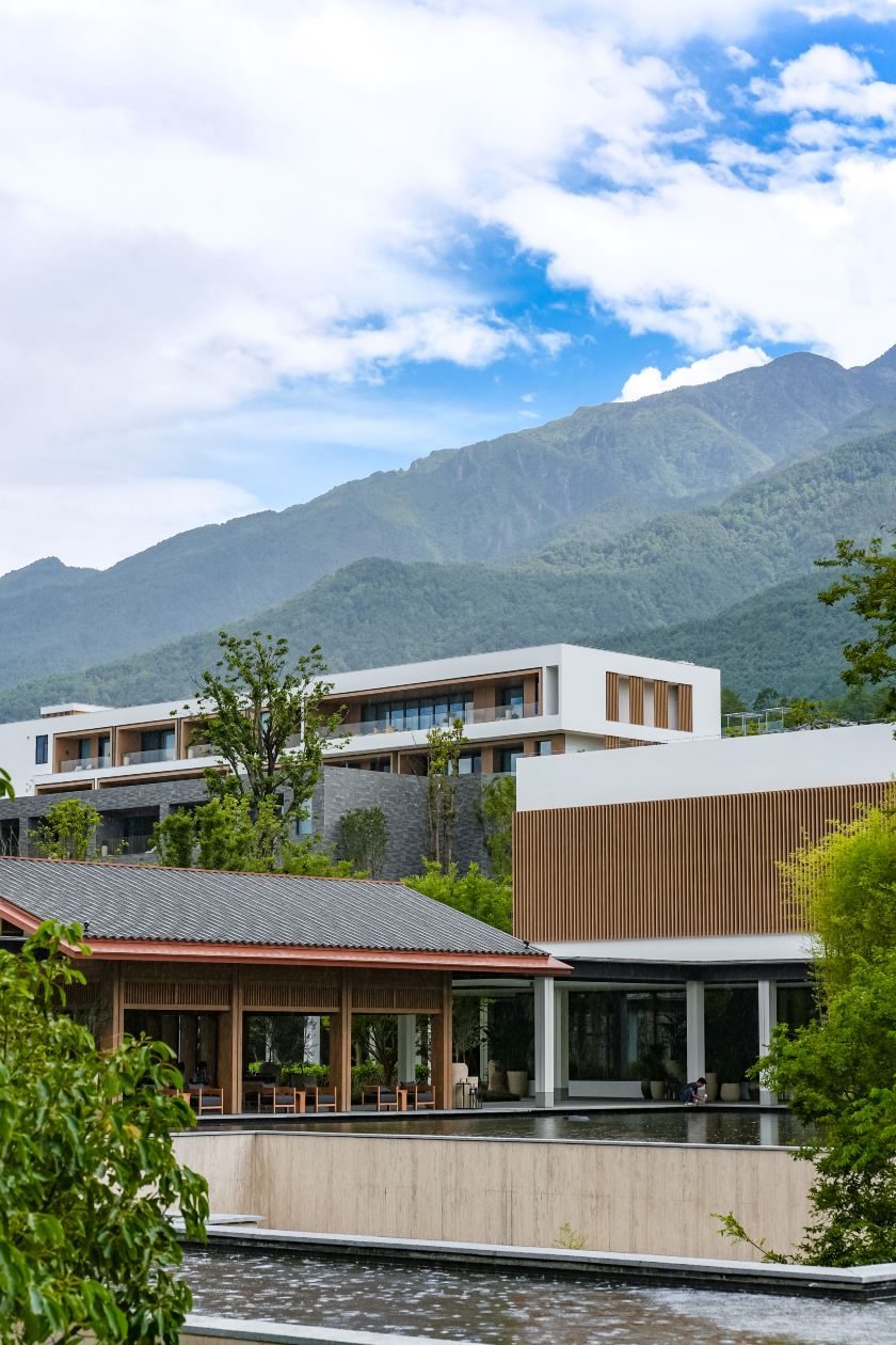 White-walled guest wings and a tiled-roof pavilion over a water garden, with the green ridges of Cangshan behind.
