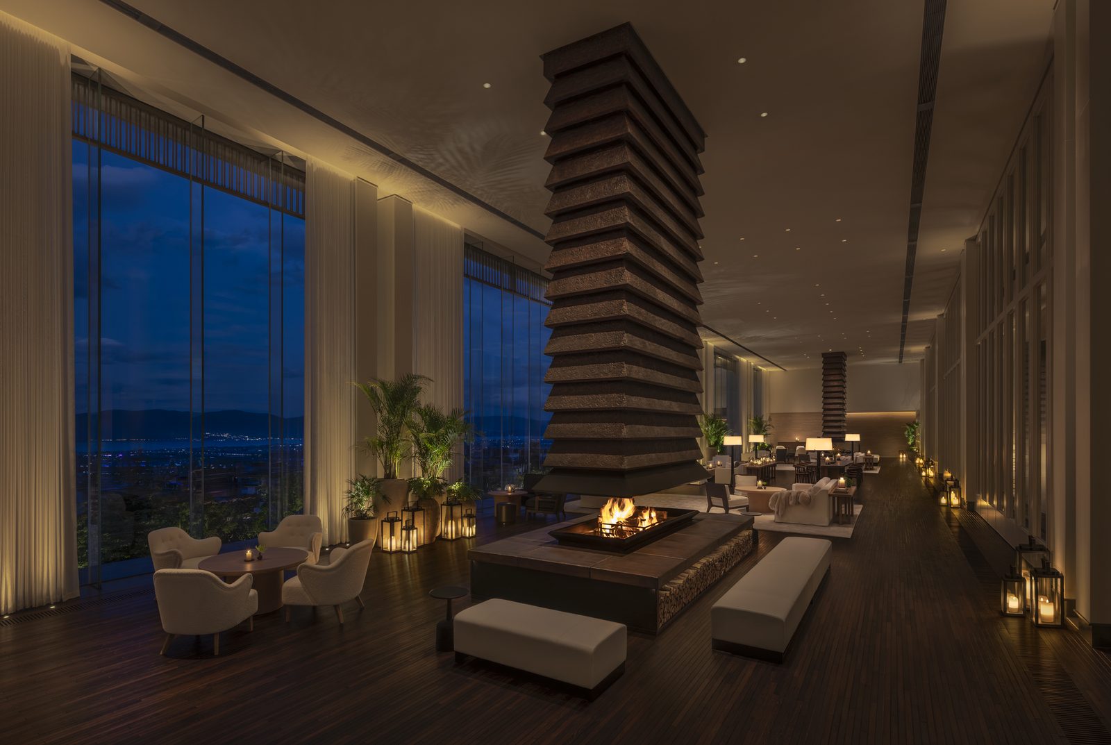 Lobby lounge at twilight, a stacked-timber fireplace column at its centre and floor-to-ceiling glass onto Erhai Lake.
